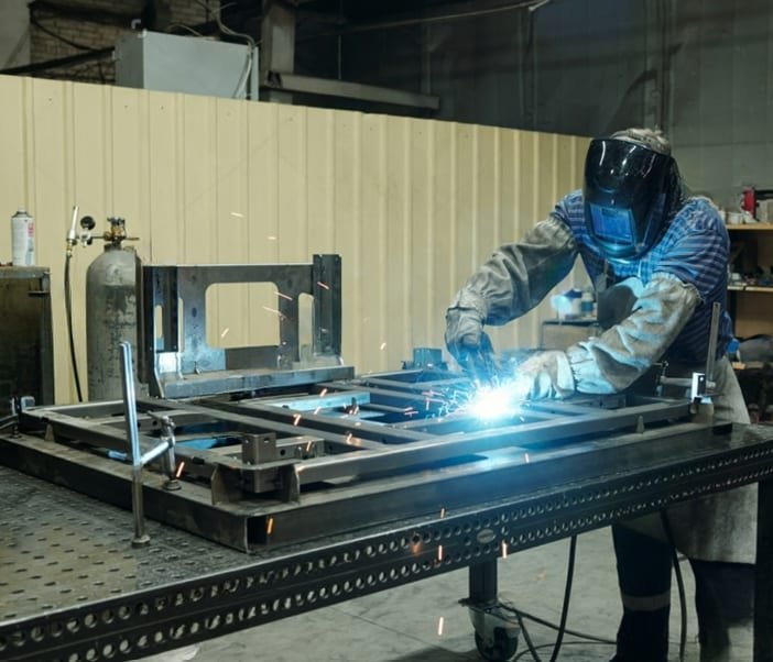 Welder in a protective helmet fabricating a steel frame on a workshop jig table